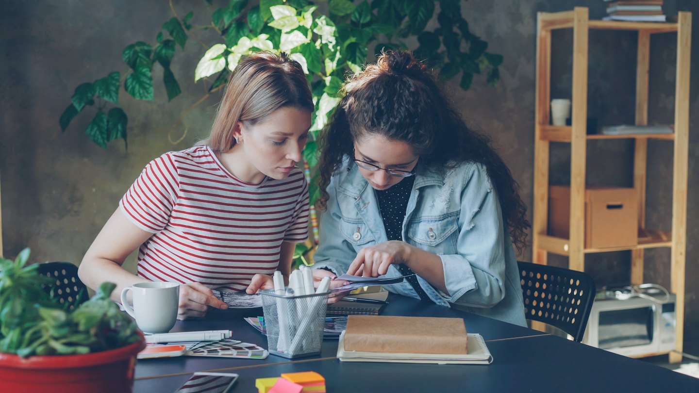 Two women collaborate over documents at a wooden table.