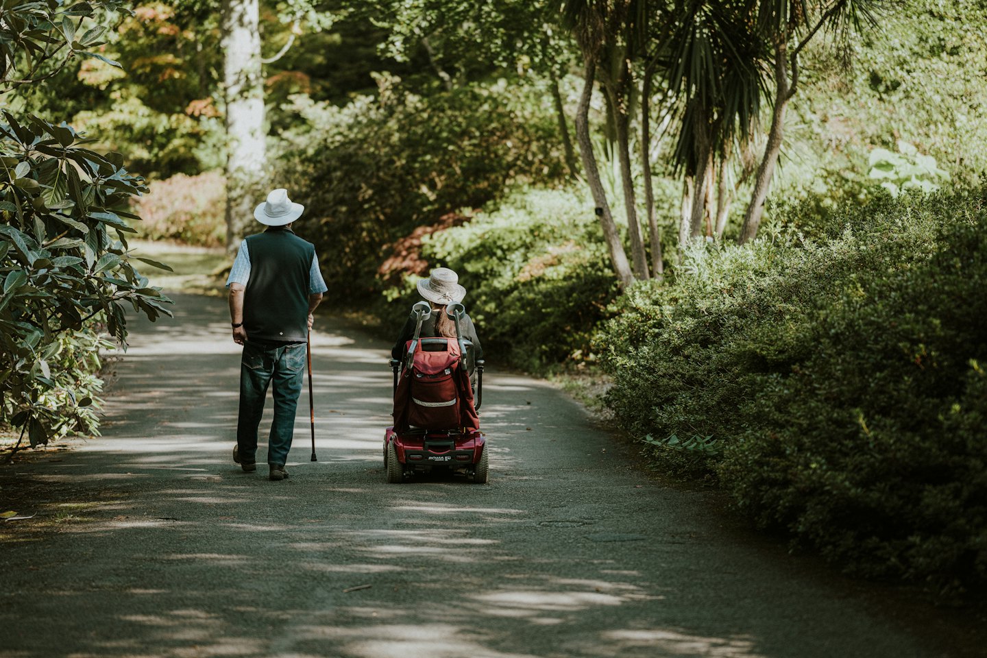 Couple walks on a sunny park path, one with a cane, the other on a scooter.